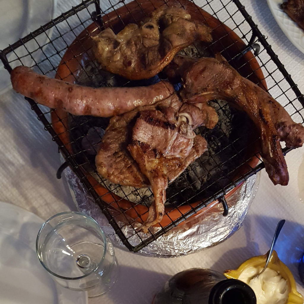 Varias piezas de carne asada (chuletas y longanizas) sobre una pequeña parrilla de mesa.