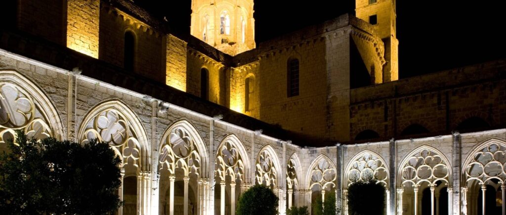 Claustro iluminado de noche, con arcos góticos y una torre del monasterio visible en la parte superior.